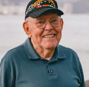 Elderly man wearing a Vietnam veteran cap, smiling near the ocean.